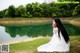 A woman in a white dress sitting on the grass by a lake.